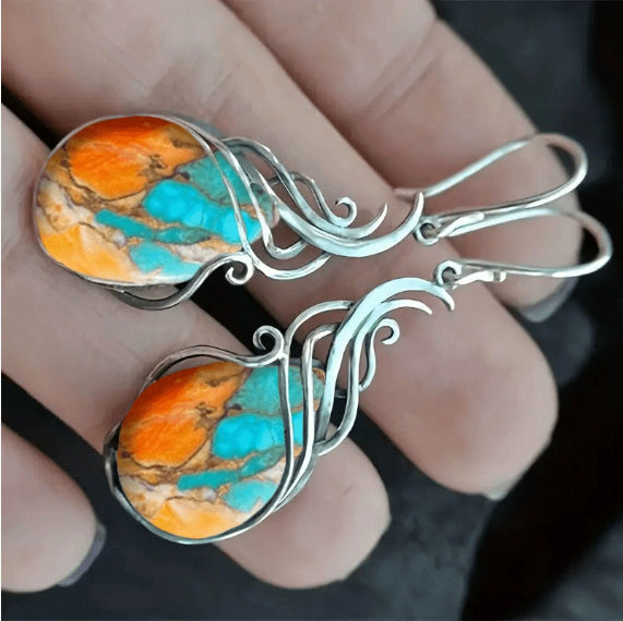 Silver earrings with orange and blue stone pendants held in a hand.