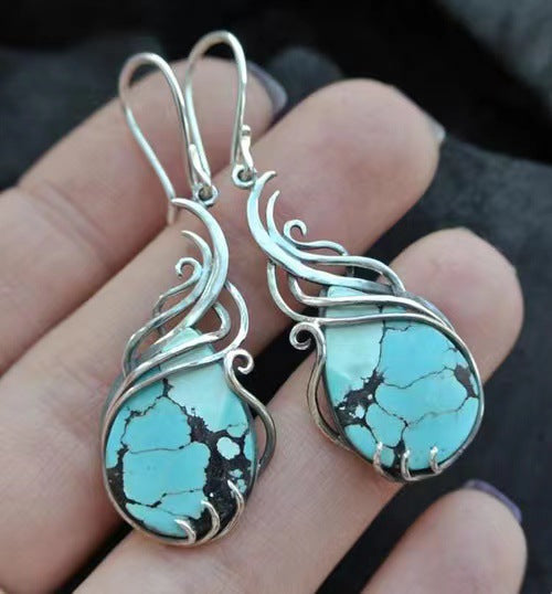 Turquoise stone earrings with silver wire wrapping held in a hand against a dark background