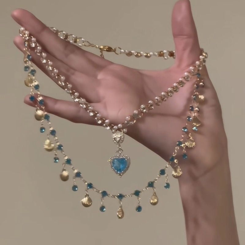 Hand holding a decorative necklace with blue heart pendant and gold accents against a neutral background