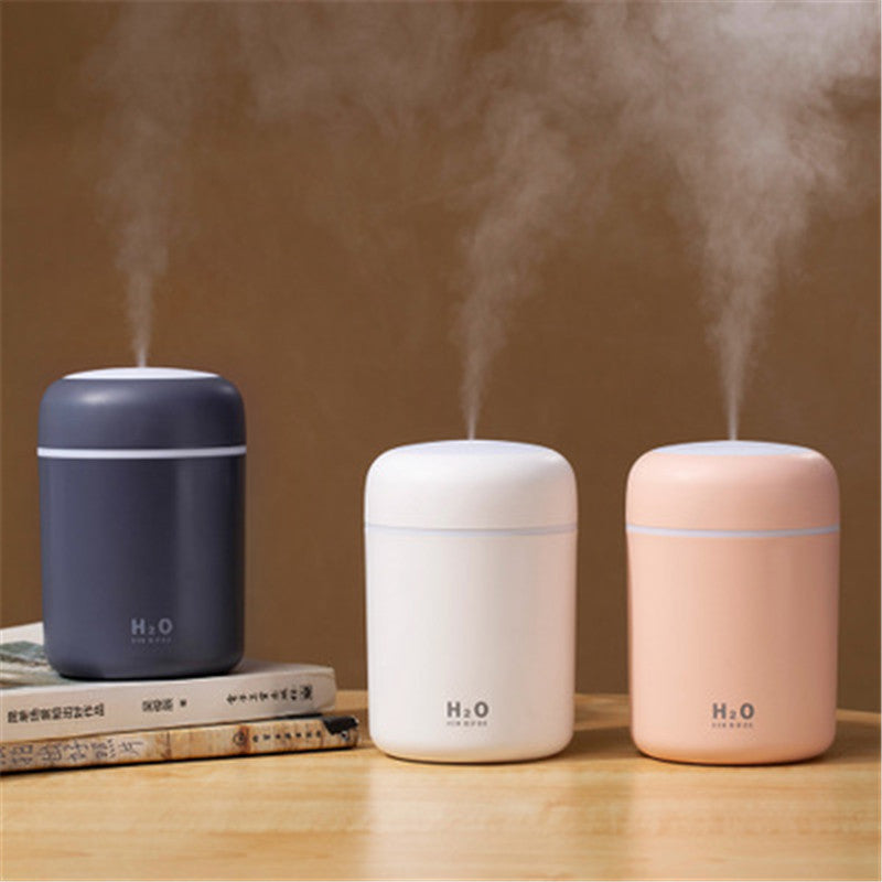 Three cylindrical humidifiers in blue, white, and pink on a wooden surface with steam rising from them.