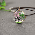 Necklace with a tree pendant featuring pink flowers and green leaves on a gray surface.