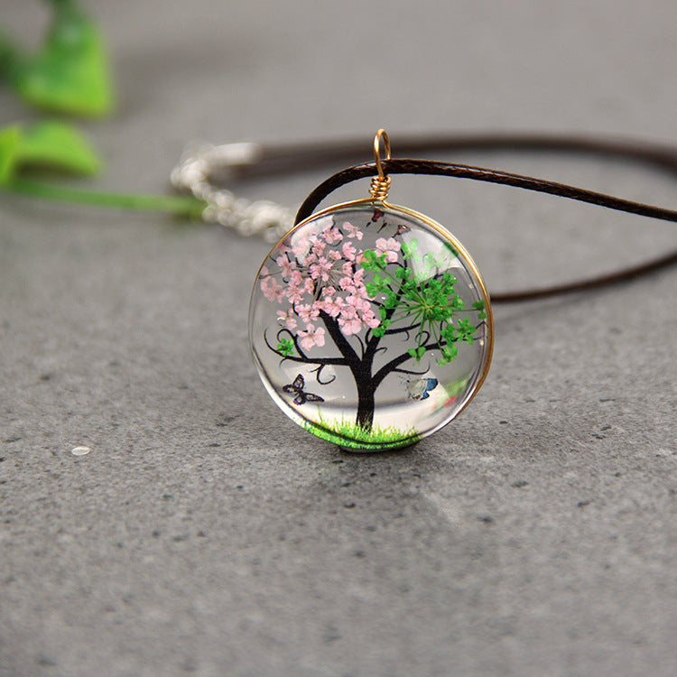 Necklace with a tree pendant featuring pink flowers and green leaves on a gray surface.