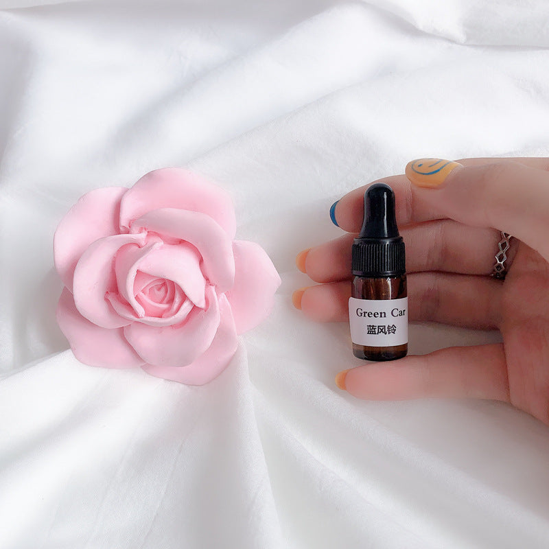 Hand holding a small bottle labeled 'Green Car' next to a pink flower on a white fabric background