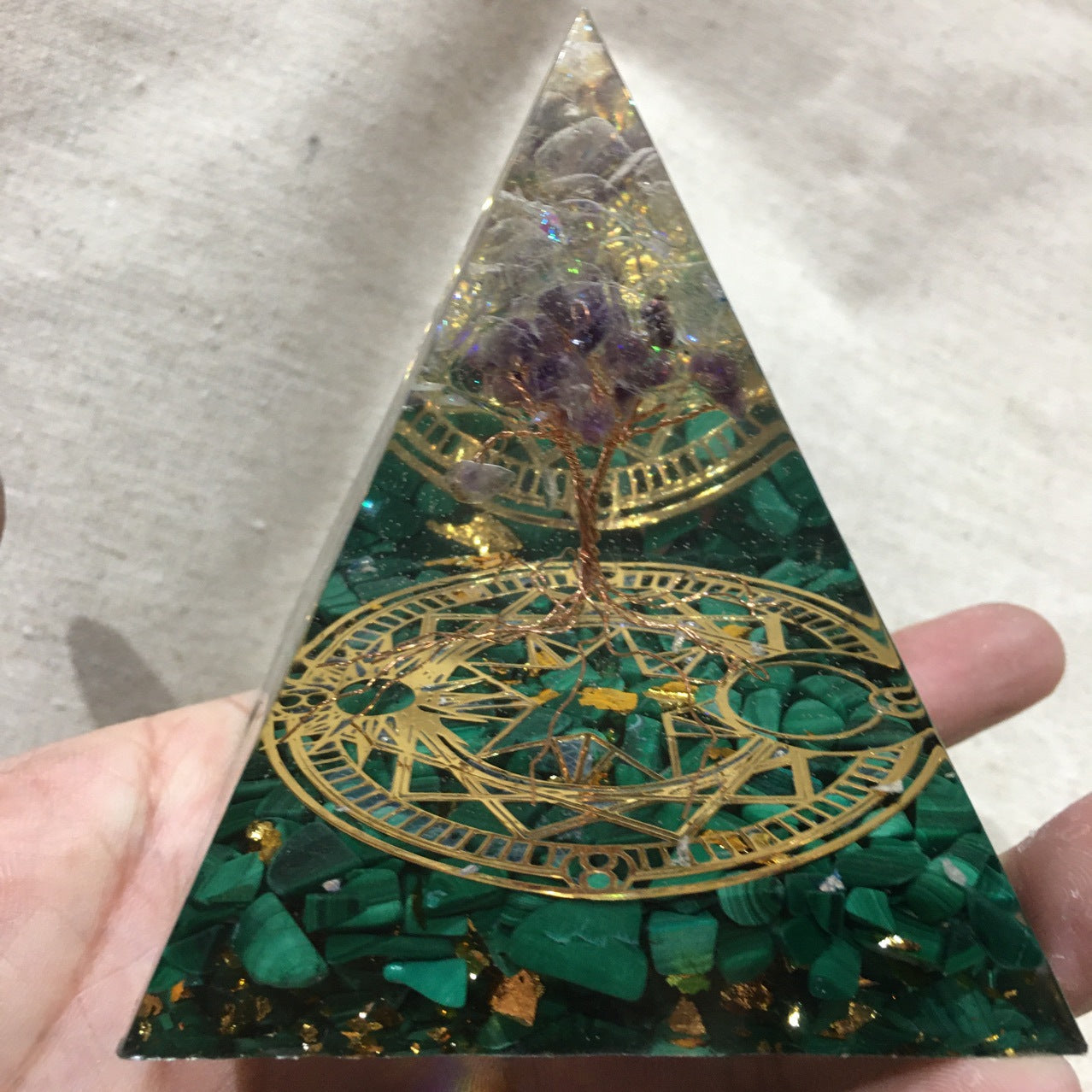 Triangular crystal with green stones and gold designs held by a hand.