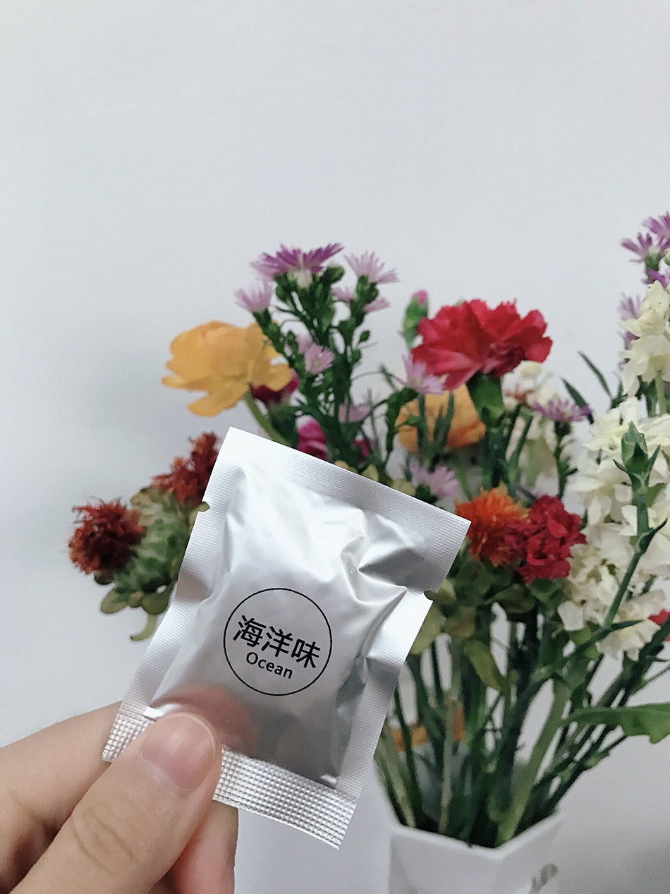 Hand holding a silver packet labeled 'Ocean' in front of a colorful flower arrangement.