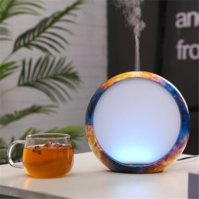 A colorful diffuser emitting steam next to a glass of tea on a surface with a blurred plant and text in the background.