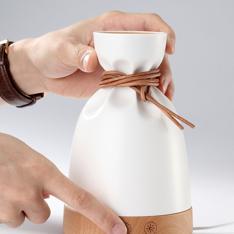 White ceramic bottle with brown rope and wooden base held by a hand on a light gray background