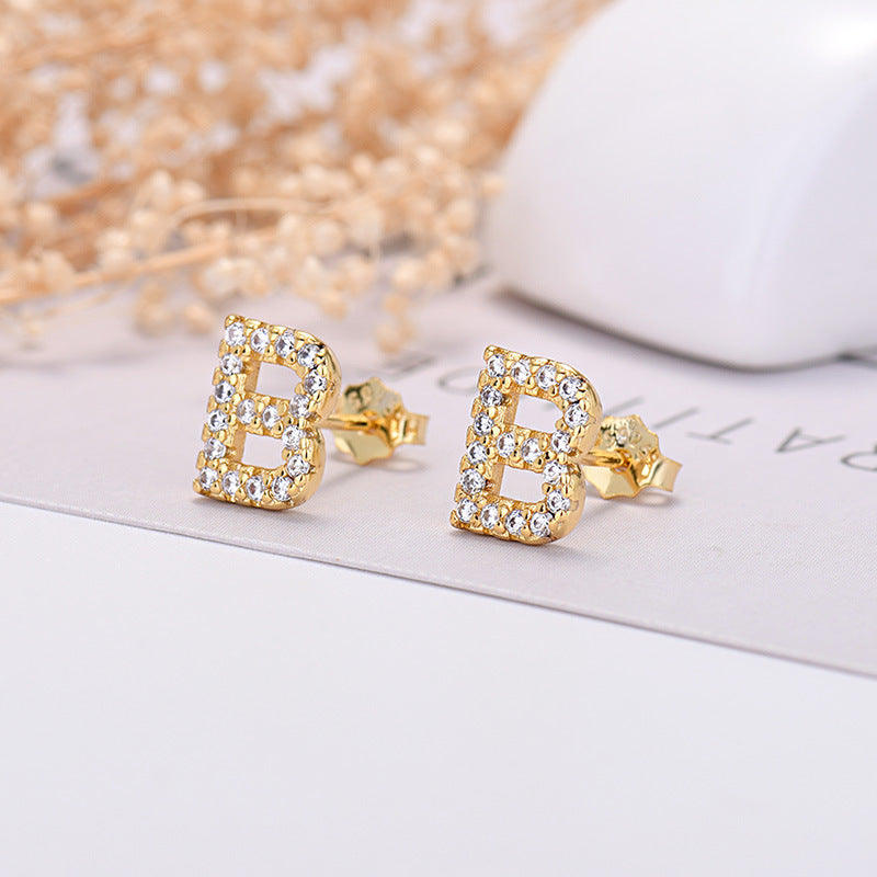 Gold letter 'B' earrings with crystals on a white surface with decorative elements.