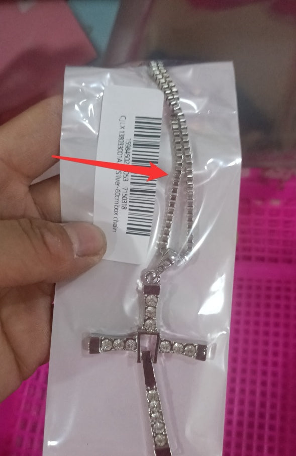 Silver cross necklace in packaging with a barcode label, held by a hand against a pink background.