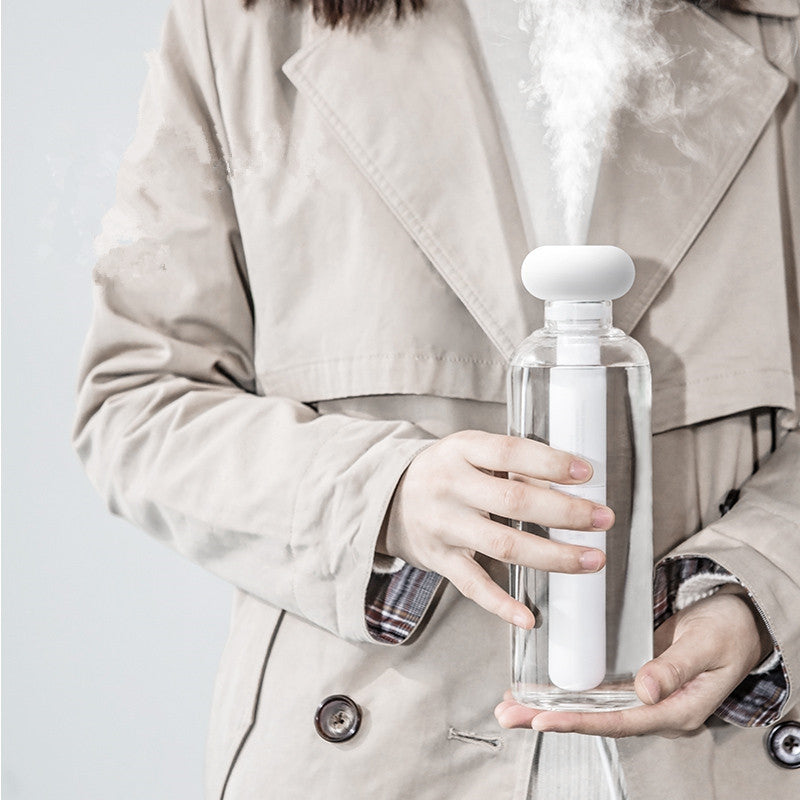 Person holding a clear spray bottle with a white nozzle, releasing mist, against a neutral background.