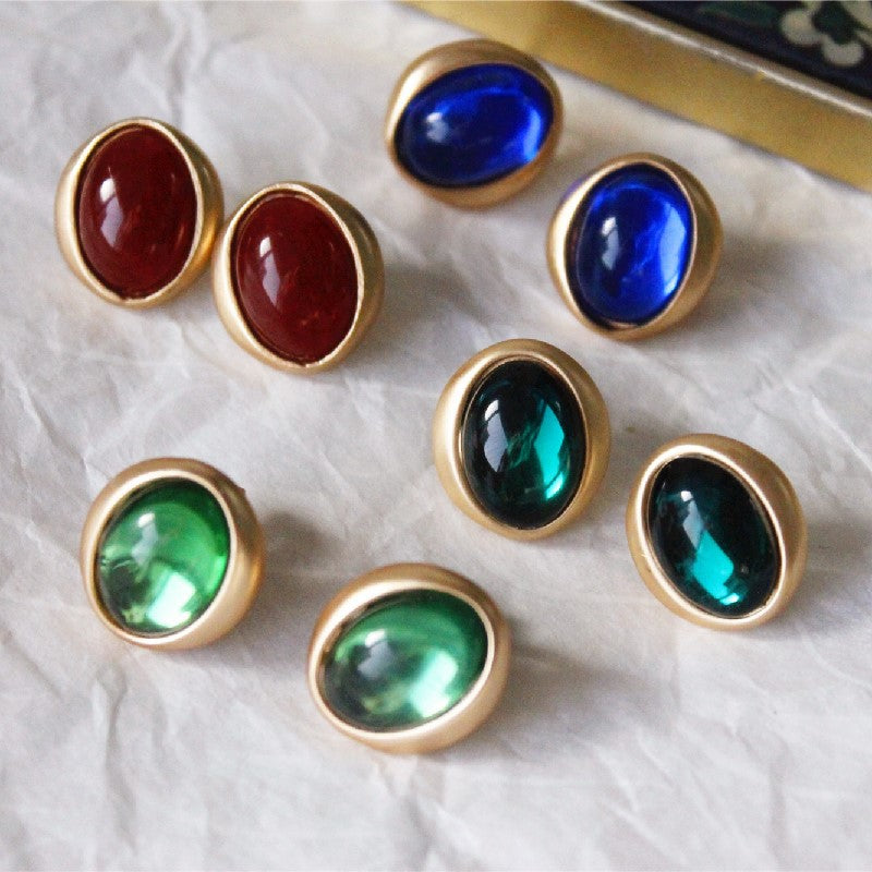 Gold-rimmed gemstone earrings with red, blue, and green stones on a textured white surface.