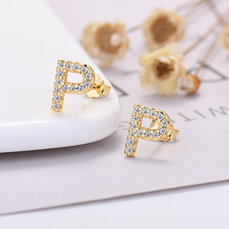 Gold earrings with letter 'P' design on a white surface with dried flowers in the background.