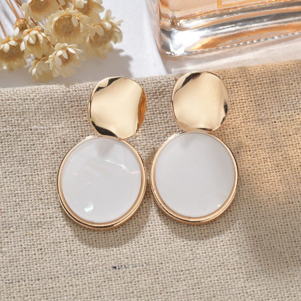 Gold and white earrings on a textured fabric surface with a bottle of perfume in the background.