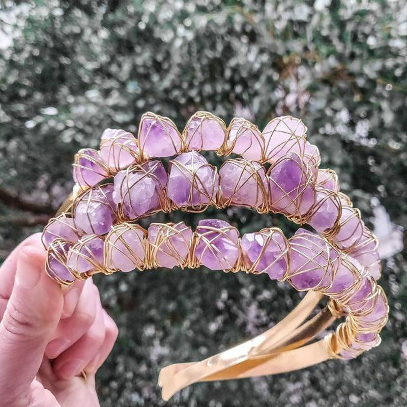 Hand holding a purple gemstone headband with gold wire wrapping against a blurred natural background