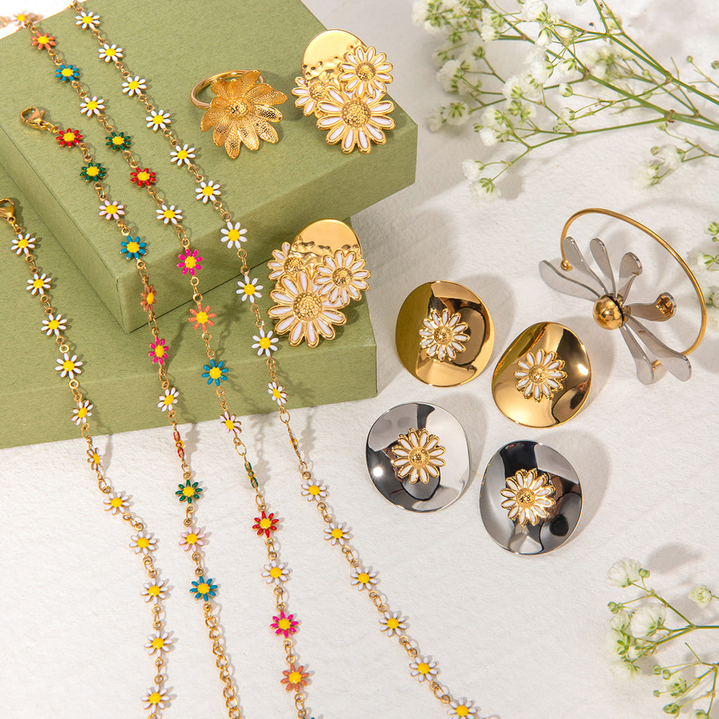 Gold jewelry set including necklaces, rings, and earrings on a white surface with green box and flowers.