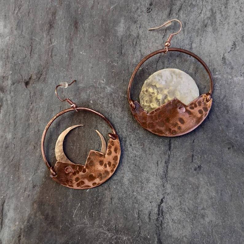 Copper earrings with moon and mountain design on a stone surface