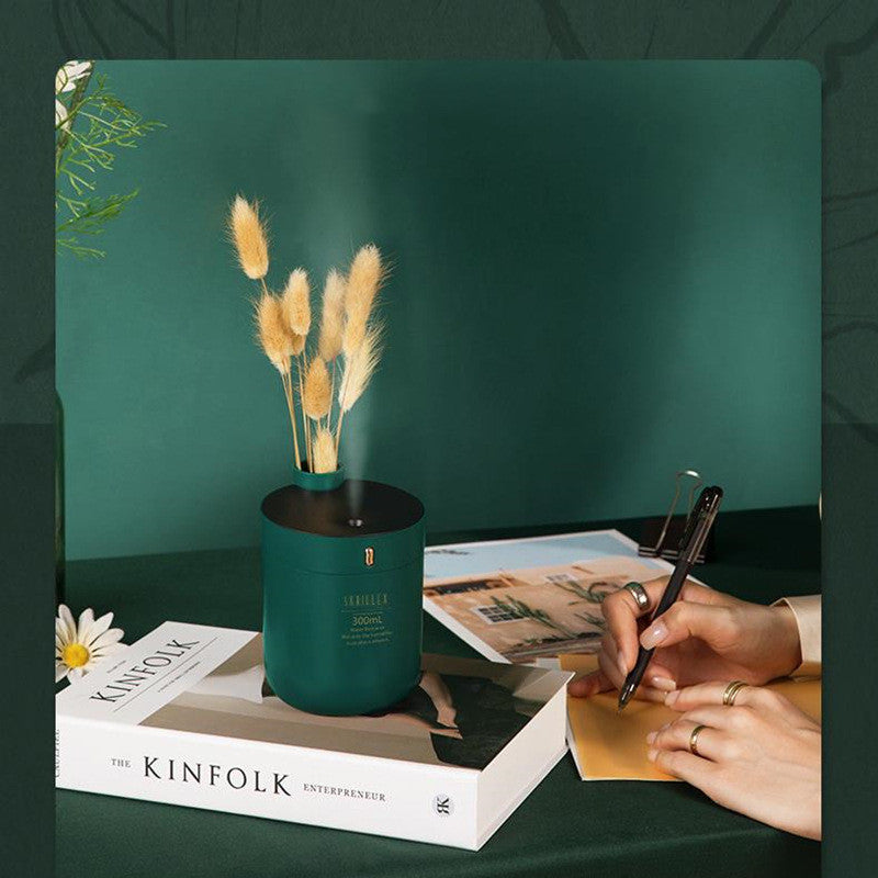 Teal diffuser with dried plants on a book, hand holding pen, against a dark green background