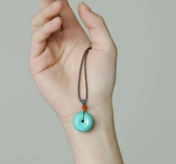 Hand wearing a necklace with a turquoise pendant on a plain background