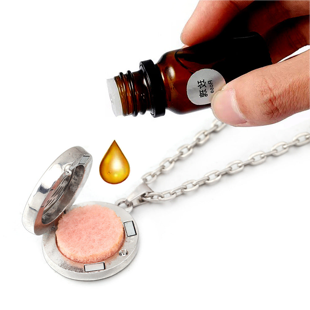 Hand pouring oil from a bottle into a silver locket with a pink stone on a white background