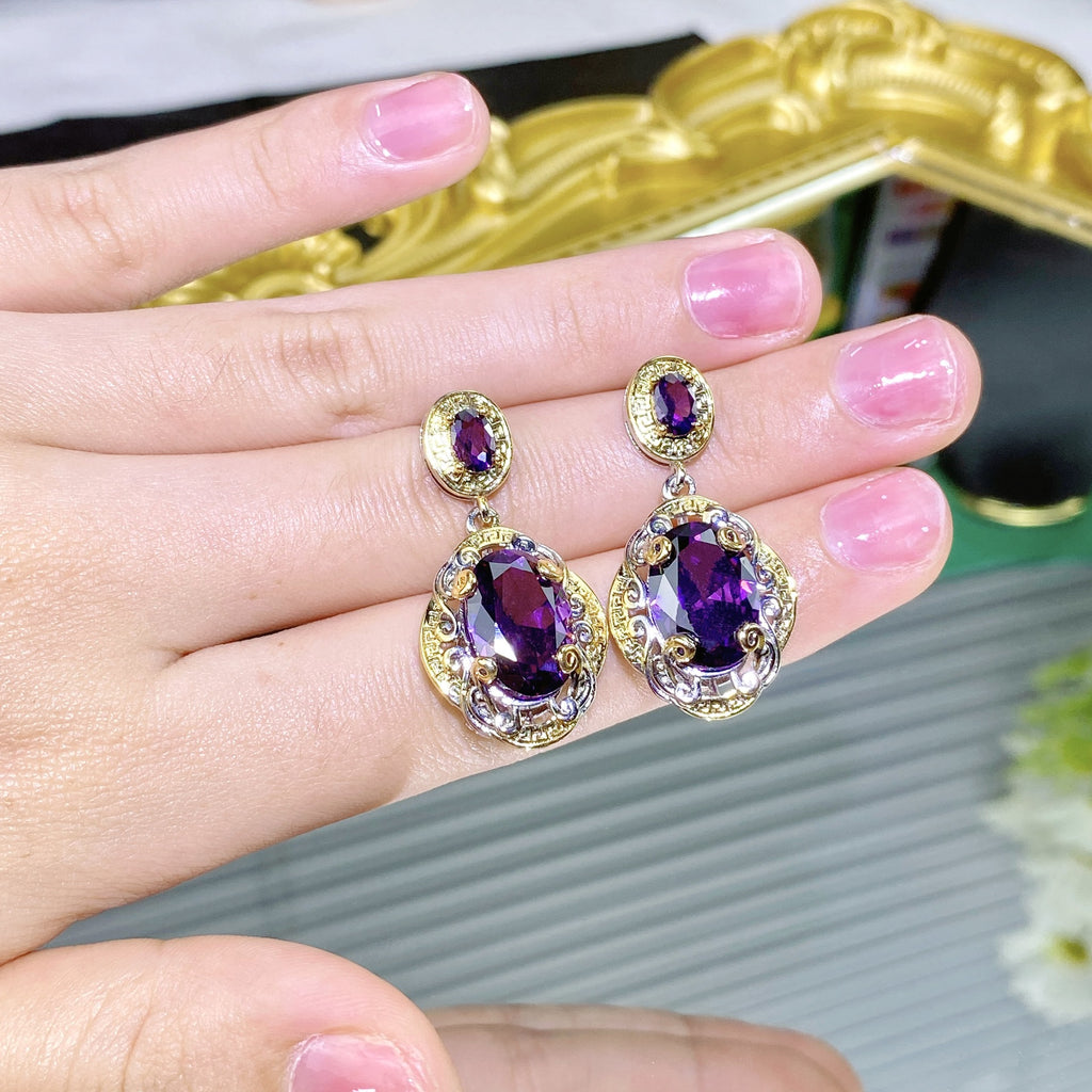 Purple earrings with gold accents held in a hand against a blurred background