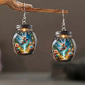 Glass jar earrings with fairy and floral design on a wooden branch against a dark background
