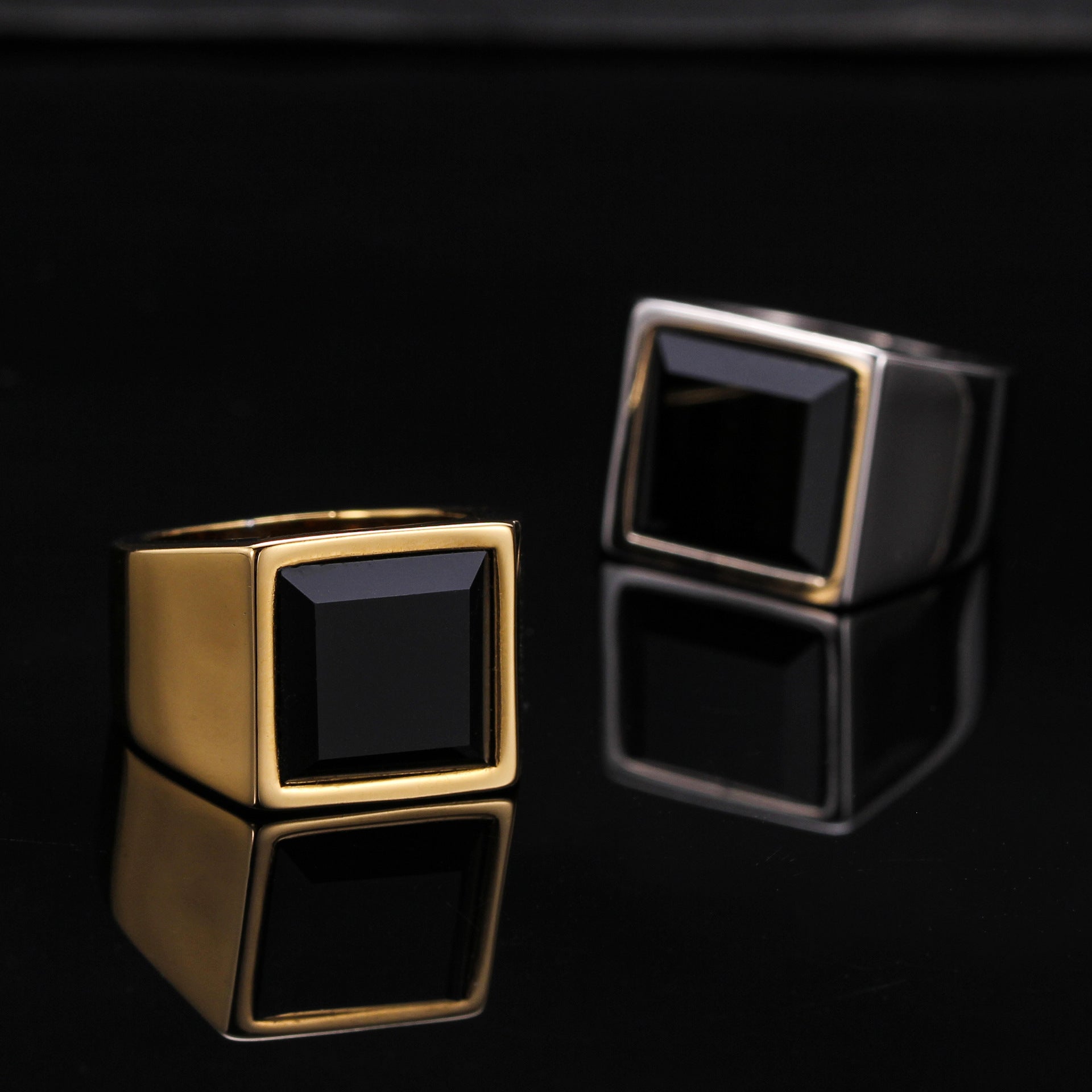 Two gold and black square ring designs on a reflective black surface.