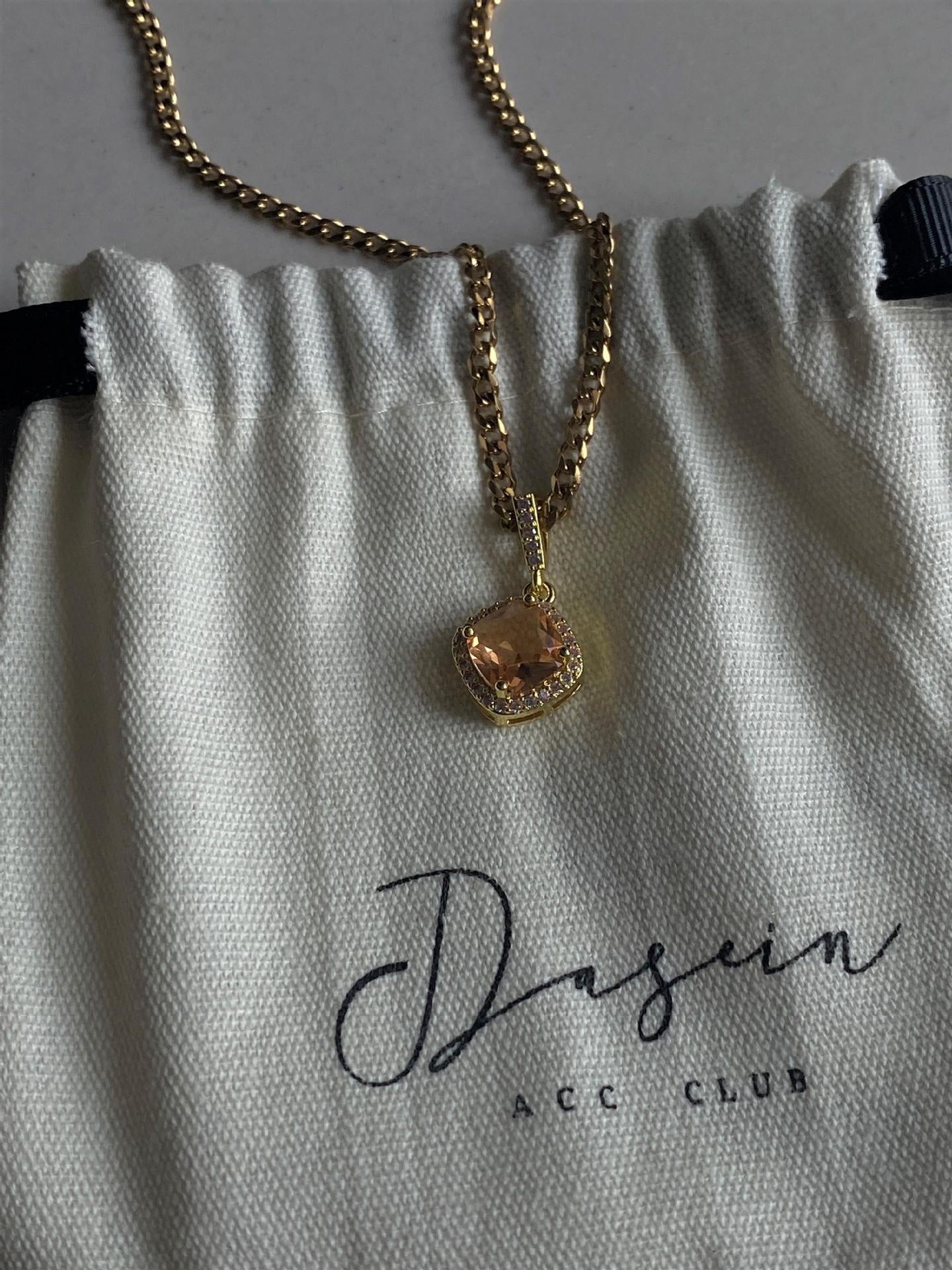 Gold necklace with a pendant on a Dasein ACC Club branded pouch.