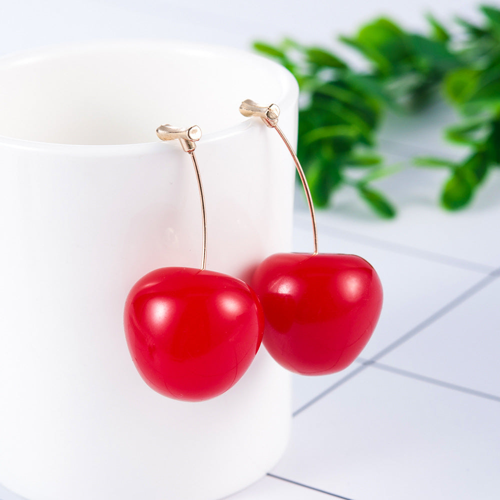 Red cherry-shaped earrings on a white surface with green leaves in the background
