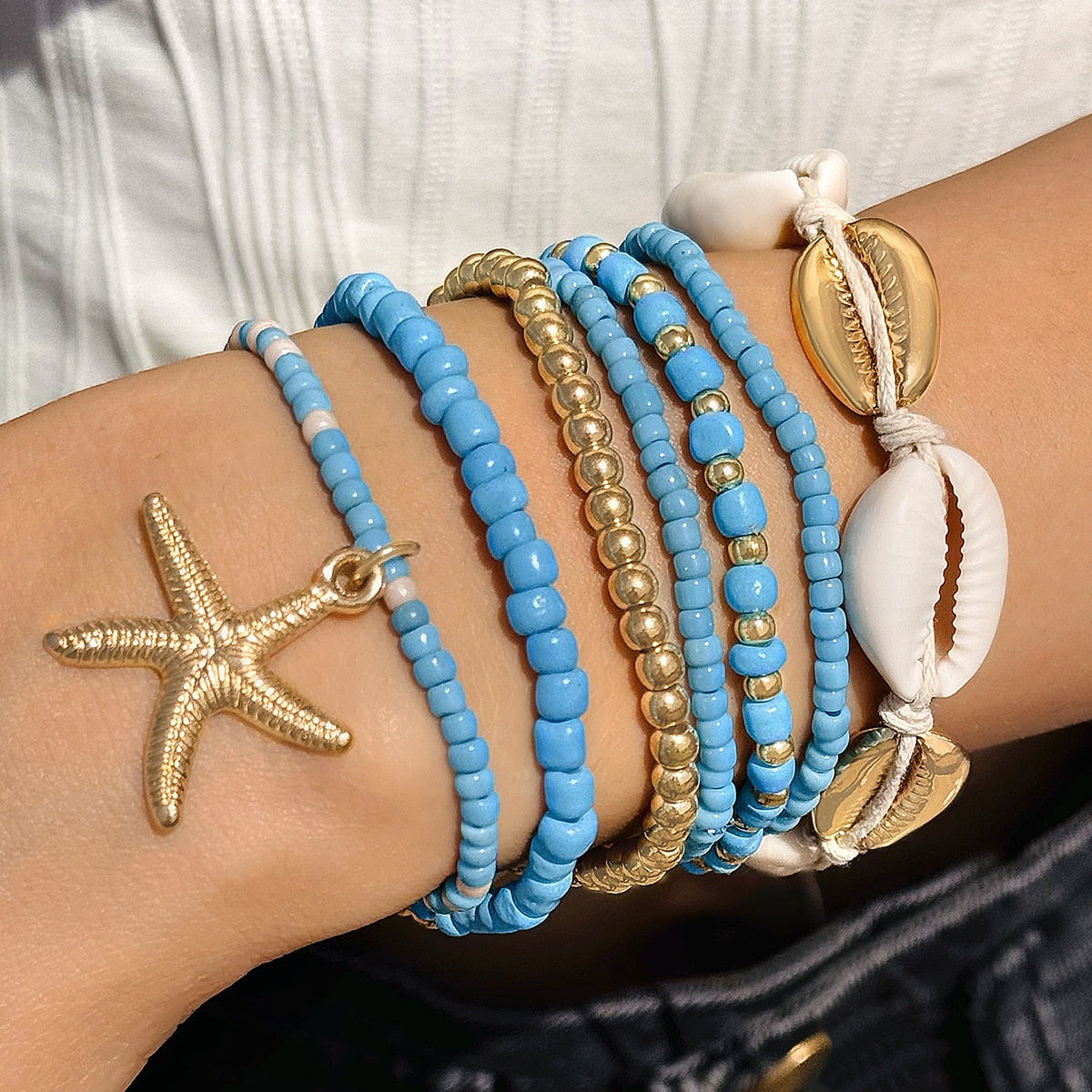 Close-up of a wrist wearing multiple blue and gold beaded bracelets with shell and starfish charms.