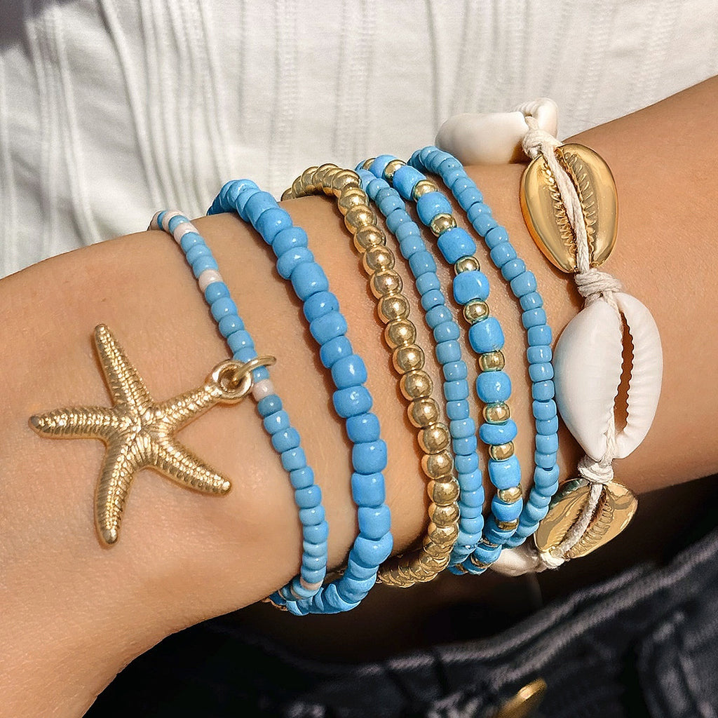 Close-up of a wrist wearing multiple blue and gold beaded bracelets with shell and starfish charms.