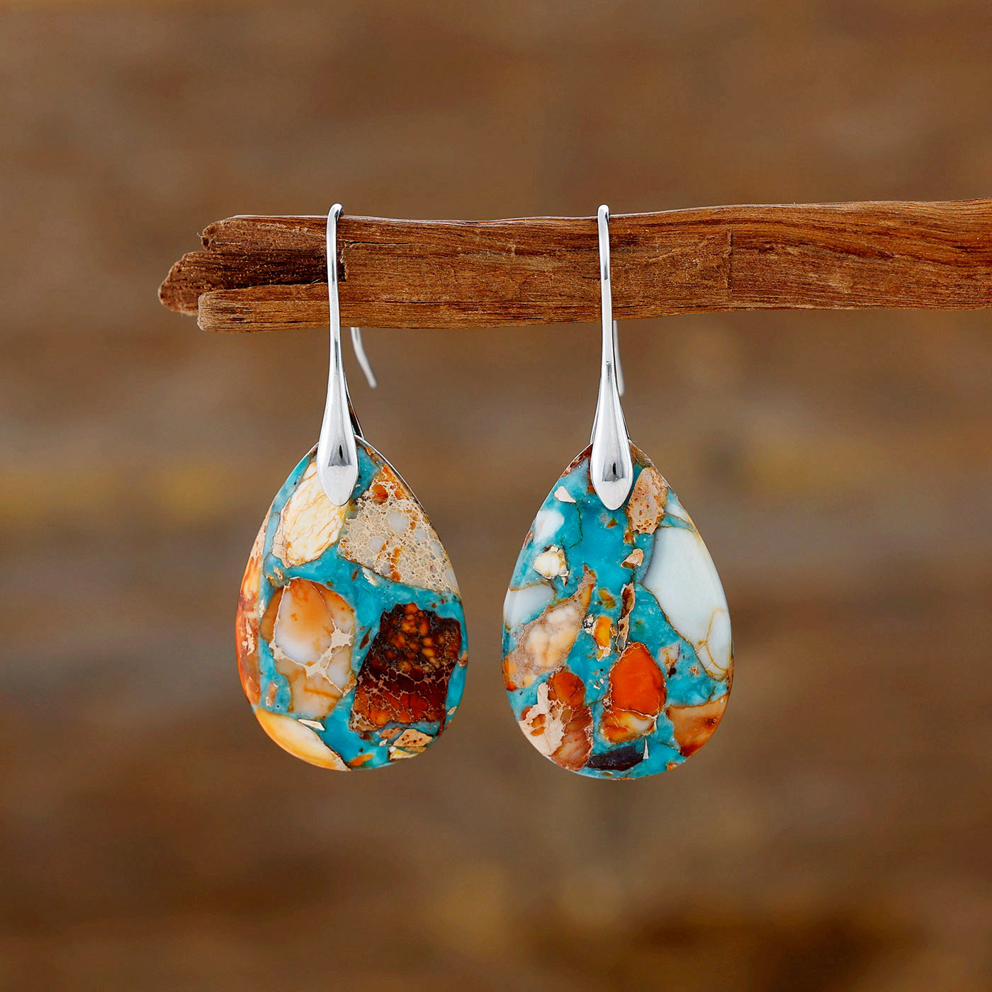 Teardrop-shaped earrings with multicolored stone design hanging on a wooden stick against a blurred natural background.