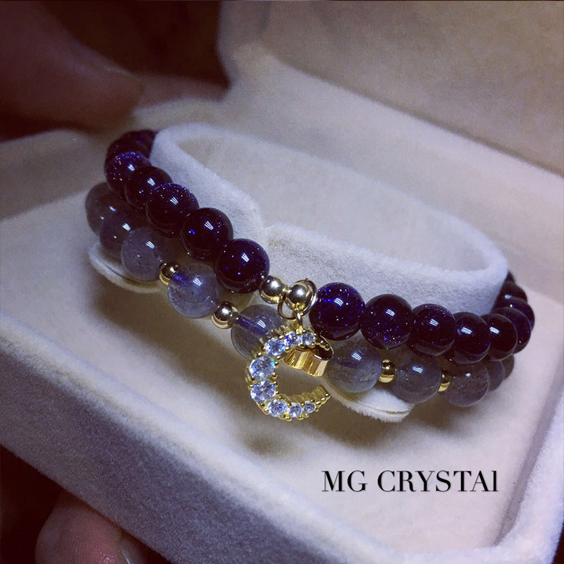 Two beaded bracelets with a gold charm on a white cushion, branded 'MG Crystai'.