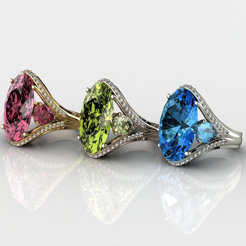 Three gemstone rings with pink, green, and blue stones on a reflective surface.