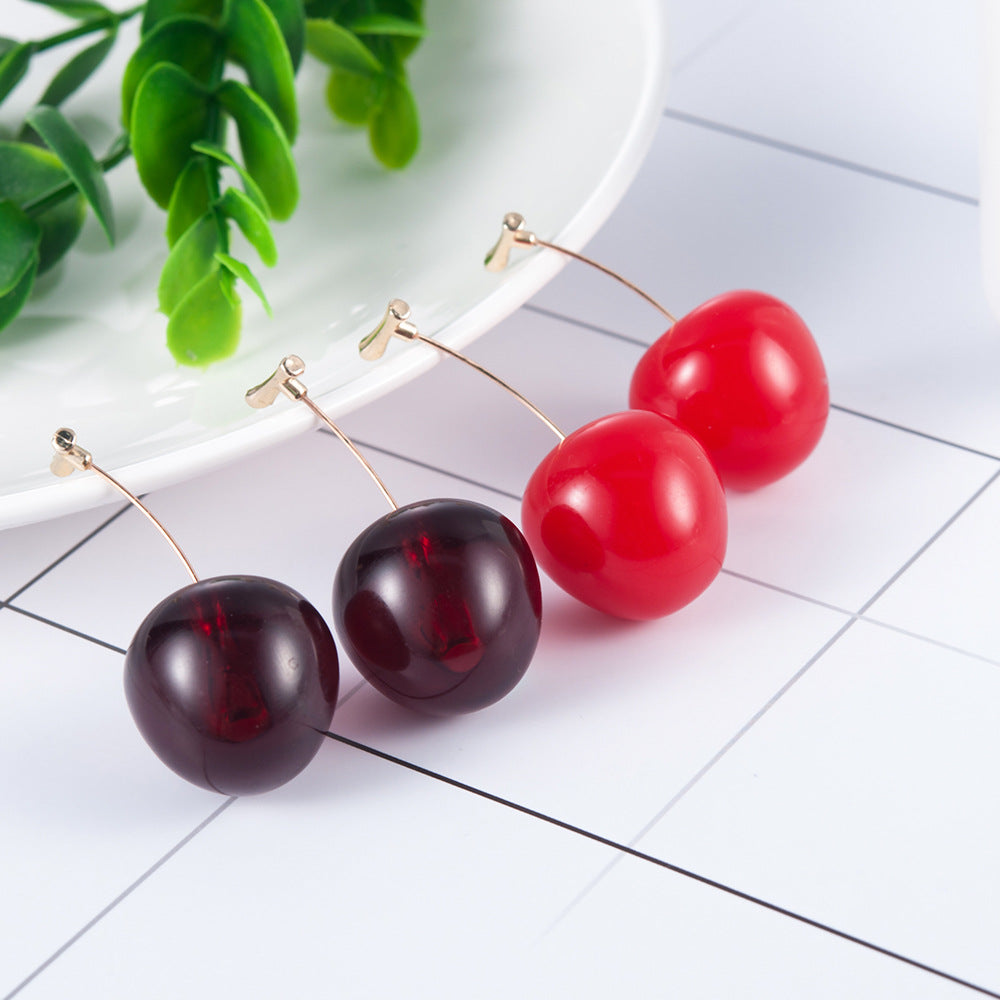Cherry-shaped earrings on a white surface with green leaves in the background