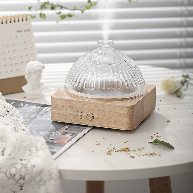 A clear glass diffuser on a wooden base emitting mist, placed on a table with decorative items.