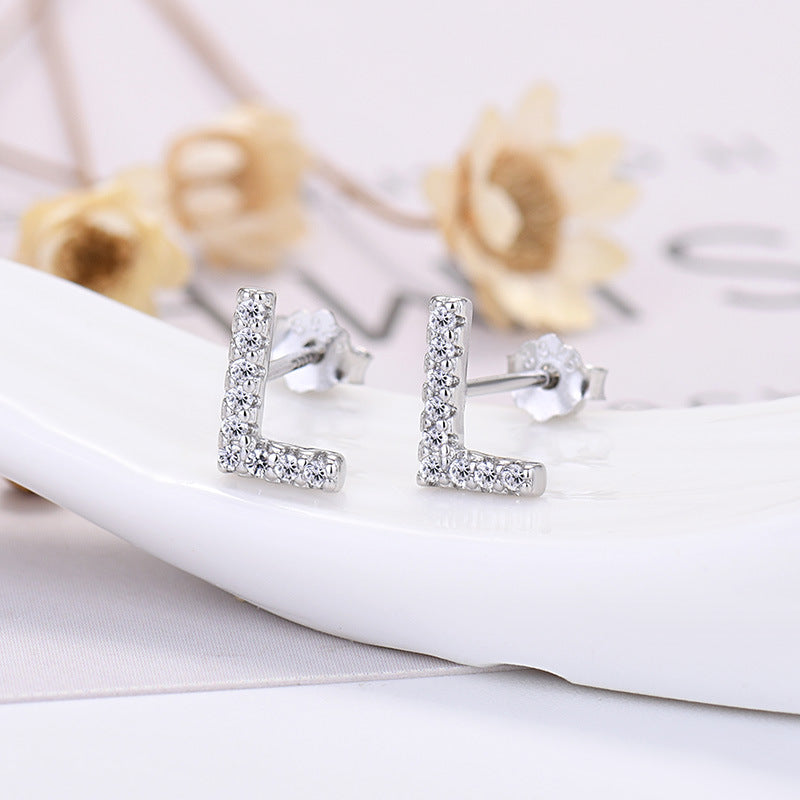 Silver 'L' shaped earrings with clear stones on a white surface with blurred flowers in the background.