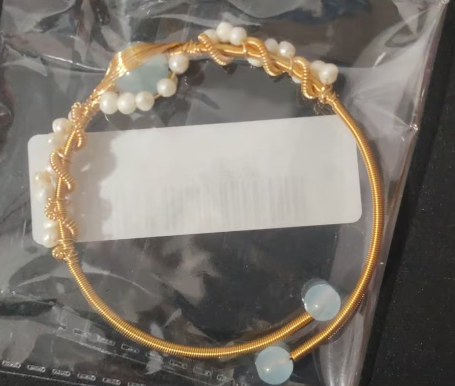 Gold bracelet with pearls and a clear stone on a white card, on a reflective surface.