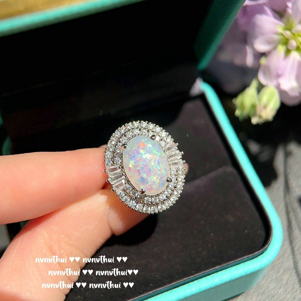Opal ring with silver setting held by a hand, blurred background with flowers.