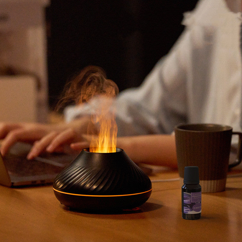 Black essential oil diffuser with flames on a wooden table, person using laptop in the background
