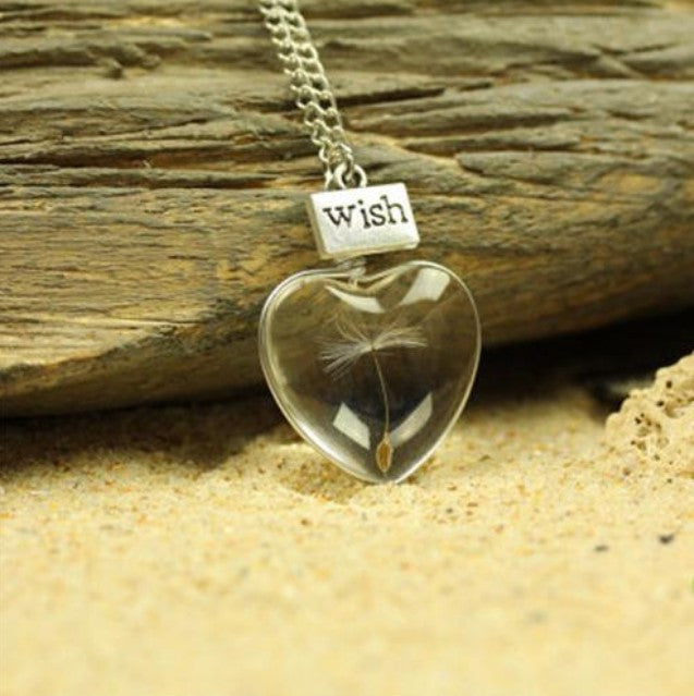 Heart-shaped pendant with a dandelion seed and 'Wish' tag on a wooden and sandy background