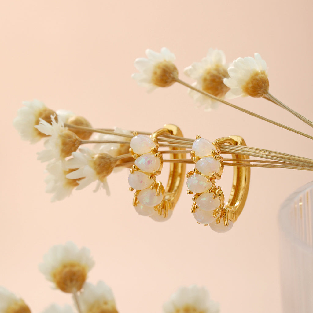 Gold hoop earrings with pearl-like stones on a branch of white flowers against a soft pink background