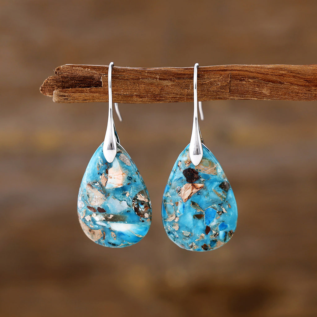 Teardrop-shaped blue earrings with embedded materials on a wooden branch against a blurred natural background.