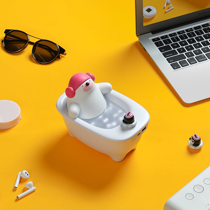 Small polar bear figurine in a bathtub with a pink hat on a yellow surface, surrounded by a laptop, sunglasses, and earbuds.