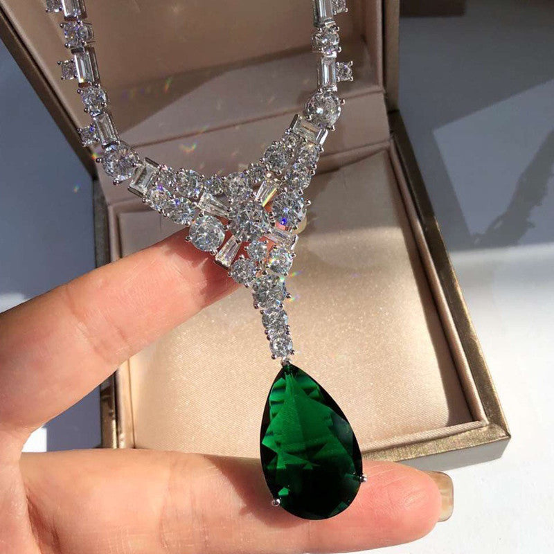Diamond necklace with a green teardrop pendant held by a hand against a beige box.