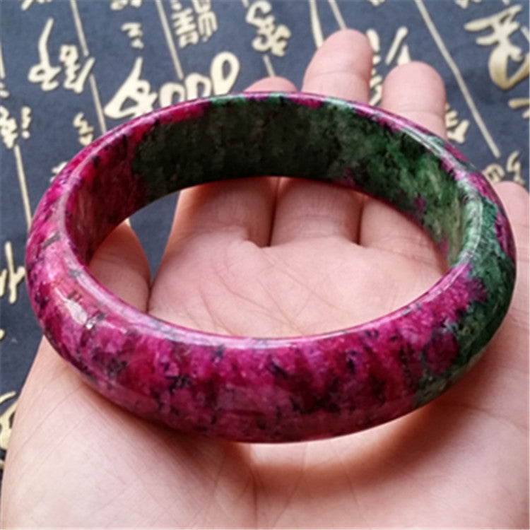 Hand holding a pink and green bangle against a dark background with gold text.