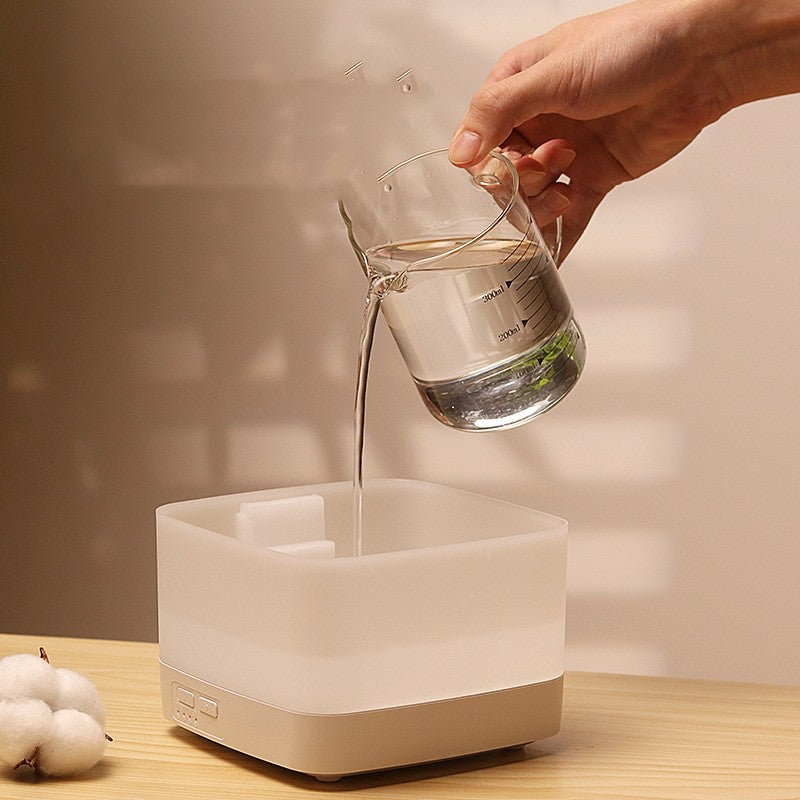 Hand pouring water from a glass into a white humidifier on a wooden surface.