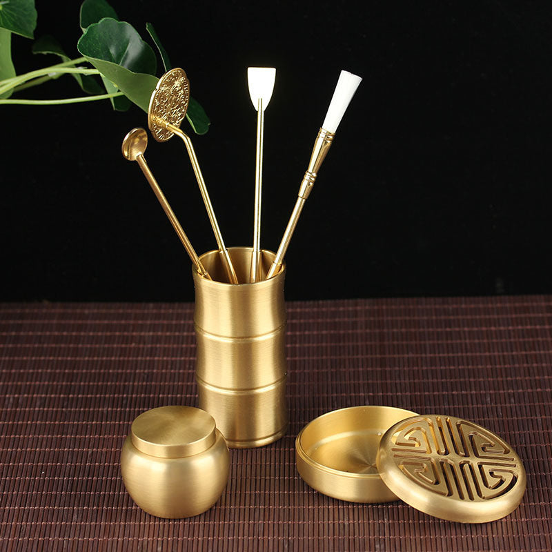 Set of gold-colored tea accessories including a container, lids, and sticks on a dark background.