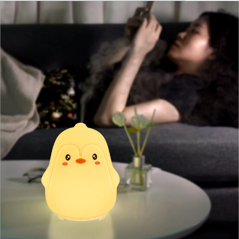 Yellow chick-shaped night light on a table with a person lying on a couch in the background.