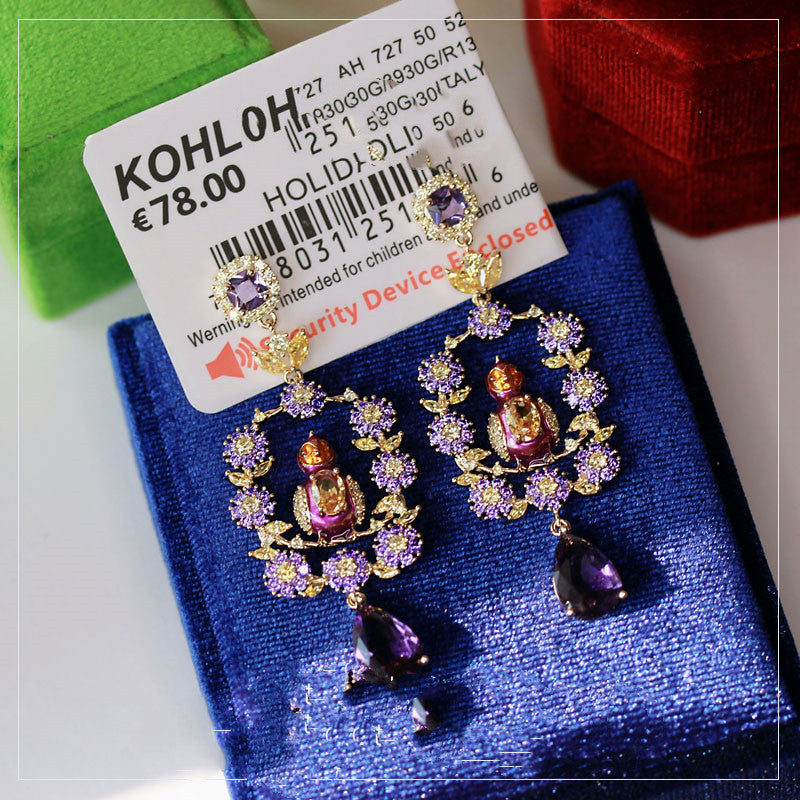 Decorative earrings with purple and gold details on a blue cushion, with a Kohloh price tag.