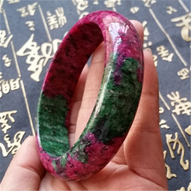 Hand holding a colorful bangle with a dark background featuring gold text.